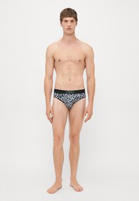 Un modèle masculin, debout pieds nus contre un fond blanc, portant des slips Versace à imprimé léopard noir et blanc avec une ceinture noire.