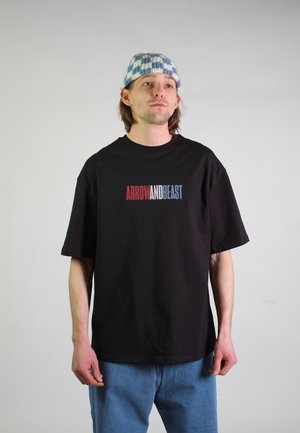 Man die een zwart T-shirt draagt met de tekst "ARROW AND BEAST" in rood, wit en blauw, blauwe spijkerbroek en een blauw-wit geruit gebreide muts.