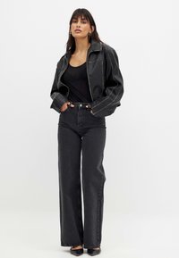 Moteris, vilkinti juodą odinę striukę ant juodo viršaus, aukštu juosmeniu, plačiomis juodomis kelnėmis ir juodais smailiais batais, stovi su rankomis kišenėse.