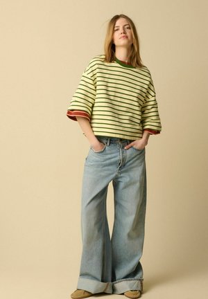 Jeune femme portant une chemise oversize à rayures vertes, un jean large avec revers et des chaussures beiges, debout les mains dans les poches devant un fond uni.