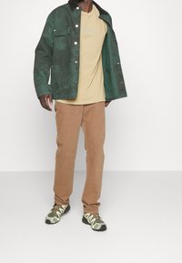 Zelená denimová bunda, béžové tričko, hnědé sametové kalhoty a tenisky s maskáčovým vzorem. Bunda má knoflíky a límec.