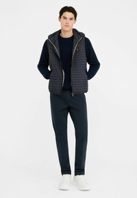 Gilet imbottito blu navy con design trapuntato verticale, cappuccio regolabile, chiusura frontale con zip, indossato sopra un maglione scuro e abbinato a pantaloni blu navy e sneakers bianche.
