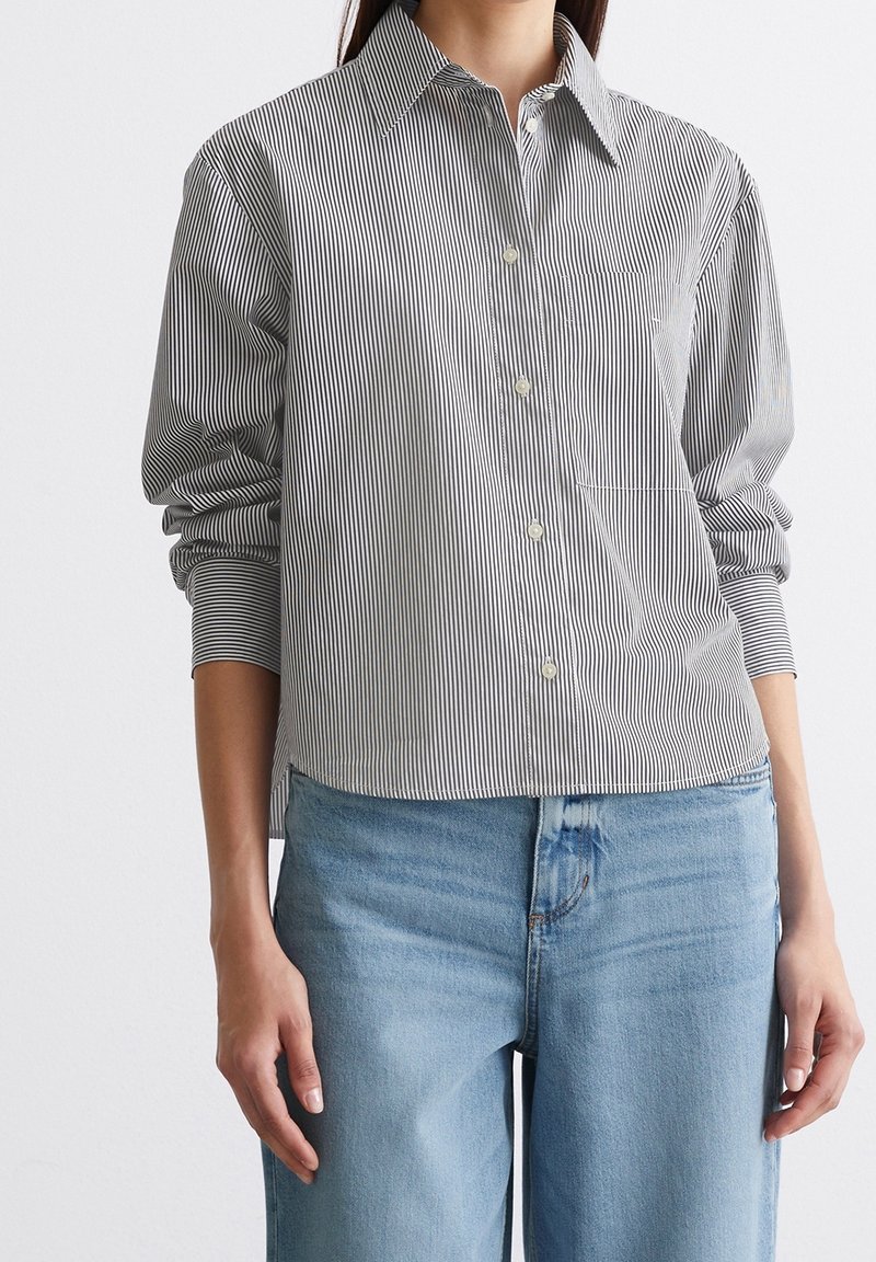 Femme portant une chemise à rayures noires et blanches à manches longues, non rentrée, sur un jean bleu clair.