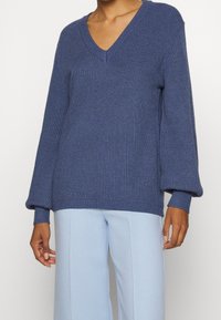 Pull bleu en maille à col en V avec larges manches et ourlet côtelés. Porté par-dessus un pantalon bleu clair, mettant en valeur la texture et la coupe ajustée.