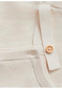 Material tricotat de culoare crem, cu un model texturat, având un nasture din lemn deschis și o curea din țesătură.