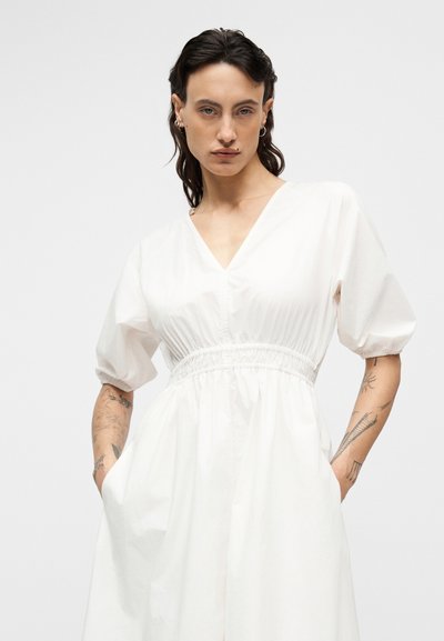 Άσπρο φόρεμα με V-λαιμόκοψη, φουσκωτά μανίκια και ελαστική μέση με πιέτες. Διαθέτει δύο πλαϊνές τσέπες και λείο, απαλό ύφασμα.