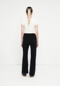Pantalons noirs évasés en tissu doux, portés avec un t-shirt blanc ajusté. Les pantalons présentent une taille côtelée et une texture subtile.