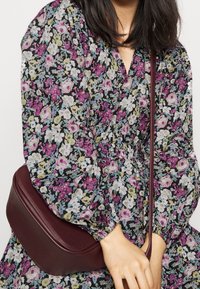 Kvinna i en blommig klänning med puffärmar som håller en vinröd läder crossbody-väska med en lång rem.