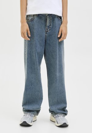 Personne portant un jean bleu ample, des baskets blanches et une chemise blanche, debout devant un fond clair uni.