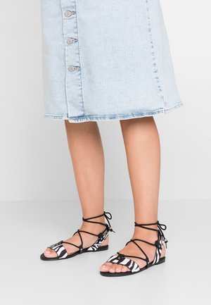 Beine mit hellblauem Denimrock und schwarz-weißen Sandalen mit Zebramuster und Knöchelriemen auf einem hellgrauen Boden.