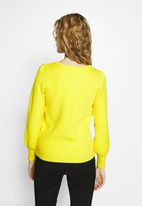 Gelbes Strickpullover mit Puffärmeln, gerippten Bündchen und Saum. Die Textur ist weich mit einem subtilen diagonalen Muster; getragen mit schwarzen Hosen.