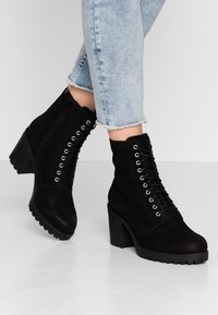Persona che indossa stivali neri in suede con lacci e tacco blocco, abbinati a jeans cropped blu chiaro, su uno sfondo bianco.