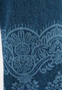 Μπλε τζιν ύφασμα με λεπτομερές σχέδιο παϊνσλεϊ σε πιο ανοιχτές μπλε αποχρώσεις, που δείχνει υφή και περίπλοκα ανθικά και σπειροειδή μοτίβα.