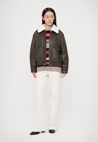 Geacă maro din piele cu guler alb din blană, două buzunare frontale, asortată cu un pulover cu dungi multicolore și pantaloni albi cu croială largă.