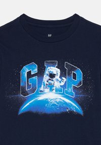 Kék ing, amelyen a "GAP" felirat csillagokkal van kitöltve, és egy lebegő űrhajós látható egy fénylő Föld felett a mellrészen.