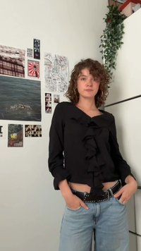 Sort bluse med volanger foran, dyb V-udskæring og lange ærmer. Kombineret med lyseblå bukser og et spændeforsynet sort bælte.