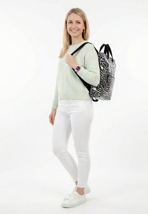 Mujer sonriente que lleva un suéter verde claro, pantalones blancos, zapatillas blancas, reloj morado, y una mochila con estampado de leopardo en blanco y negro colgada de un hombro.