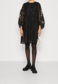 Robe noire en tissu transparent avec un motif floral, manches ballon et taille froncée. Associée à des collants noirs et des chaussures noires épaisses.