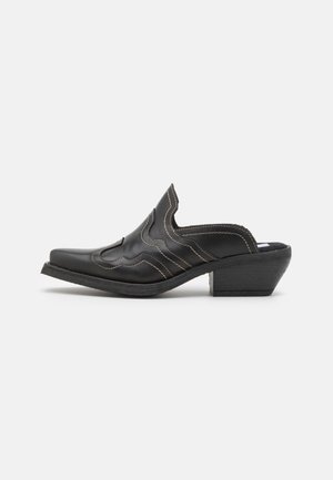 Mule en cuir noir avec bout pointu, dessus festonné avec coutures blanches, et talon empilé incliné.