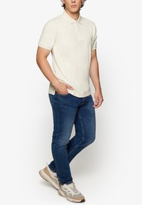 Lys beige polo shirt med klassisk krave, parret med blå denimjeans og lyse sneakers, der viser et afslappet, tætsiddende design.