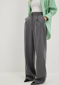 Gray high-waisted trousers with pleats, made of textured fabric. Features a wide leg design and slanted side pockets, paired with a green striped shirt.