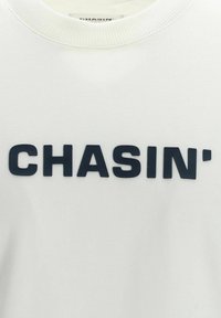 Weißes Sweatshirt mit einem gerippten Rundhalsausschnitt und erhabenen schwarzen Buchstaben, die "CHASIN'" über die Vorderseite schreiben. Weiche Stofftextur.