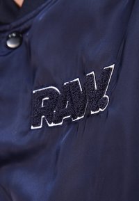Námořnická modrá bomber bunda s hladkou texturou, na které je na levé části hrudi vyšitý plyšový logotyp "RAW." v bílé a černé barvě.