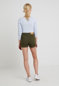 Pantalones cortos verde oliva de diseño ajustado, con un bolsillo trasero y una etiqueta de cuero marrón en la cintura, combinados con una camiseta de manga larga azul claro.