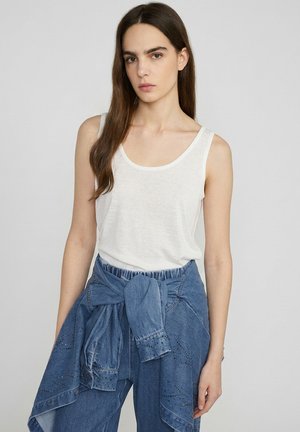 Kobieta z długimi brązowymi włosami, ubrana w biały tank top i niebieską kurtkę dżinsową zawiązaną wokół talii, stoi na tle jednolitego tła.