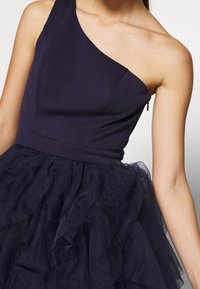 Epäsymmetrinen laivastonsininen mekko, jossa on vartalonmyötäinen yläosa ja röyhelöinen tyllihame, sileä kangaspinta ja sivulla sijaitseva vetoketjuyksityiskohta.