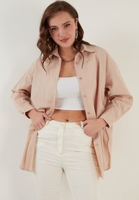 Svijetlosmeđi oversized sako s mekanom teksturom, zatvoren na dugmad, dužih rukava, uparen s bijelim rebrastim crop topom i hlačama od vrhnja visokog struka.