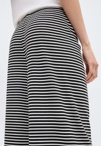 Person iført sort-hvide vandrette stribede bukser og en hvid top, stående mod en ensfarvet hvid baggrund.