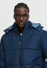 Mörkblå puffjacka med hög krage, dragkedja och justerbar dragsko på huvan. Har sydda paneler och knappdetaljer.