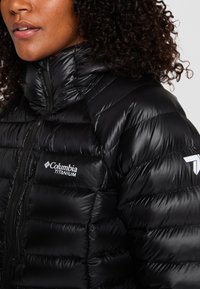 Svart isolerad pufferjacka med hög krage, med en framdragkedja, sydda detaljer och Columbia Titanium-logotyp på bröstet.
