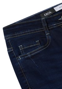 Jean denim bleu foncé avec une vue rapprochée de la ceinture, présentant une petite poche avant et des coutures contrastantes jaunes. Étiquette de la marque visible.
