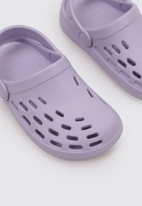 Une paire de sabots en lavande à enfiler avec des trous de ventilation et des sangles de talon ajustables sur une surface blanche.