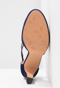 Escarpin slingback bleu marine à bout pointu, montrant une semelle beige avec un motif floral embossé et un petit talon noir.