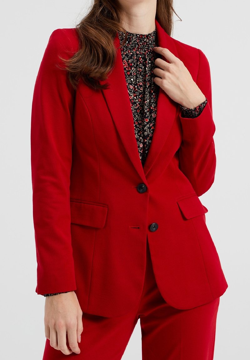 Femme portant un costume rouge avec un chemisier noir à motif floral, ajustant le col de sa veste d'une main, face à un fond uni.