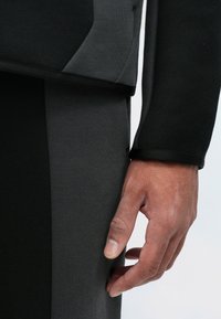 Czarny i szary warstwowy strój z fakturowanej tkaniny. Zbliżenie pokazuje ramię spoczywające na ubraniu, podkreślając szwy i dopasowanie.