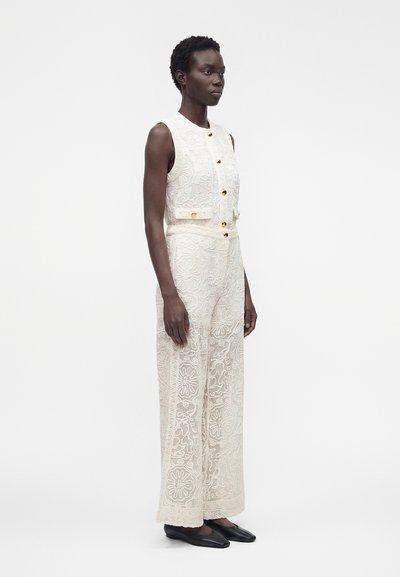 Femme debout, portant une combinaison blanche en dentelle sans manches avec des boutons dorés et des chaussures plates noires, sur un fond blanc uni.