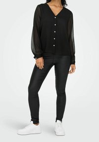 Blouse noire avec manches longues transparentes, encolure en V et fermeture à boutons sur le devant. Associée à des leggings noirs et des baskets blanches.