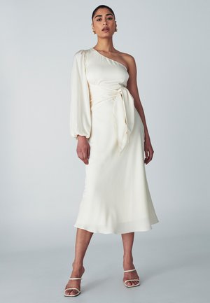 White satin midi dress with one long puff sleeve, a tied waist detail, and a subtle flowing hem. Worn with strappy white heels.