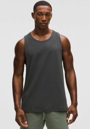 Fitness-Mann trägt ein ärmelloses dunkelgraues Tanktop und hellgrüne Shorts, steht vor einem schlichten weißen Hintergrund.