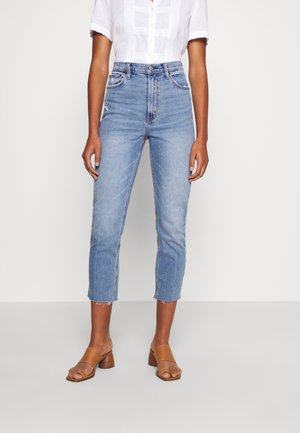 Woman wearing high-waisted, cropped light blue jeans with frayed hems, a white short-sleeve button-up shirt, and brown open-toe heeled sandals.