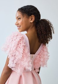 Vaaleanpunainen mekko, jossa on kukkakuviolliset puffihihat, teksturoitu kangas, istuva yläosa ja levenevä helma. Mekossa on takana vetoketjusuljenta ja kukkakoristelut hihoissa.