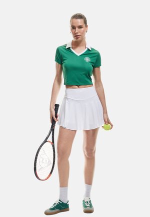 Grøn kortærmet polo skjorte med hvid krave, hvid flared tennis nederdel, grønne og hvide sneakers og et tennisracket der holder en gul bold.