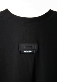 Camiseta de algodón negra con cuello redondo acanalado y una etiqueta de marca rectangular con texto en relieve y costuras en blanco contrastantes.
