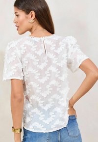 Witte blouse van lichtgewicht stof, met een textuurpatroon van bloemen, korte pofmouwen en een kleine opening met sleutelgat aan de achterkant.