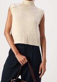 Suéter sin mangas de punto crema con cuello alto y dobladillo acanalado, combinado con pantalones oscuros. Mano sosteniendo un bolso marrón.