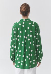 Žalia ilgomis rankovėmis marškinėliai su apykakle, puošta didelėmis baltomis taškelėmis. Lygus audinys, laisvo silueto ir su sagomis užsegami rankogaliai.